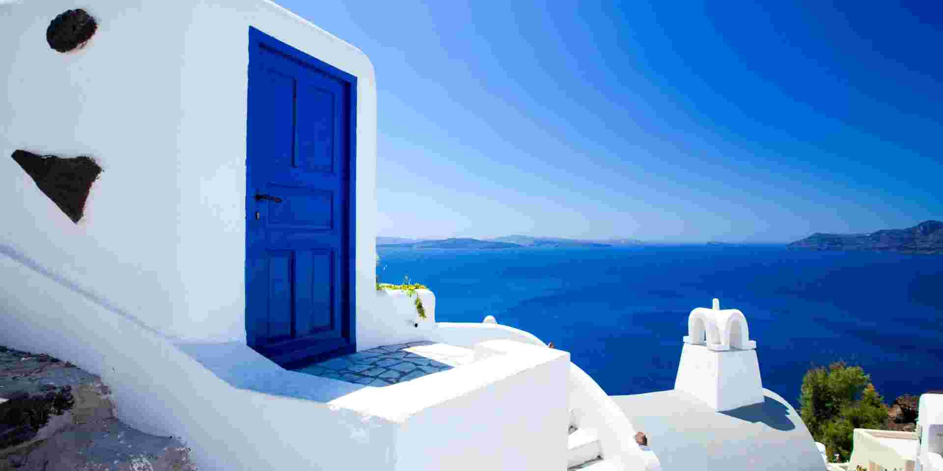 A striking blue door on a whitewashed building in Santorini, with a clear view of the deep blue Aegean Sea and distant islands under a bright, cloudless sky. reasons to visit santorini_feature
