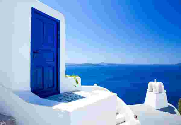A striking blue door on a whitewashed building in Santorini, with a clear view of the deep blue Aegean Sea and distant islands under a bright, cloudless sky. reasons to visit santorini_feature