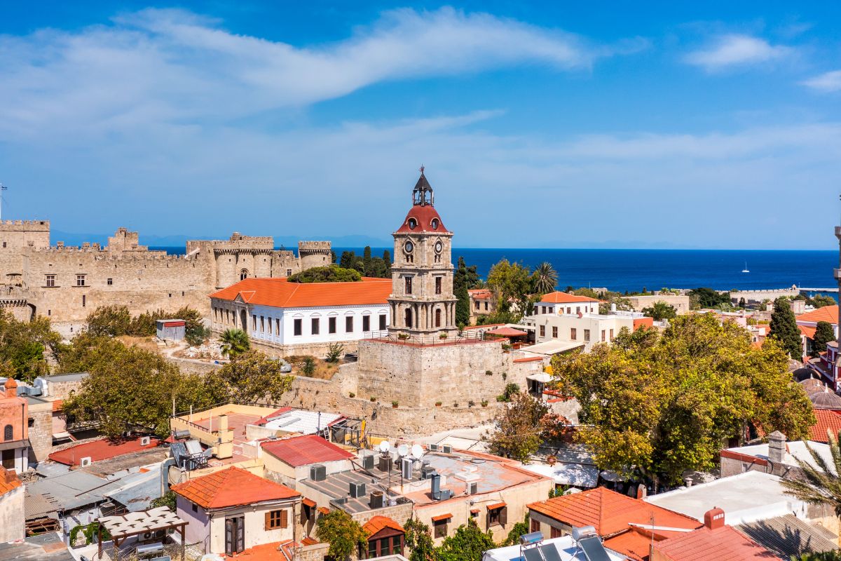 A scenic view of the old town in Rhodes, showcasing a clock tower, historic stone buildings, and the medieval castle, with the blue sea in the background.