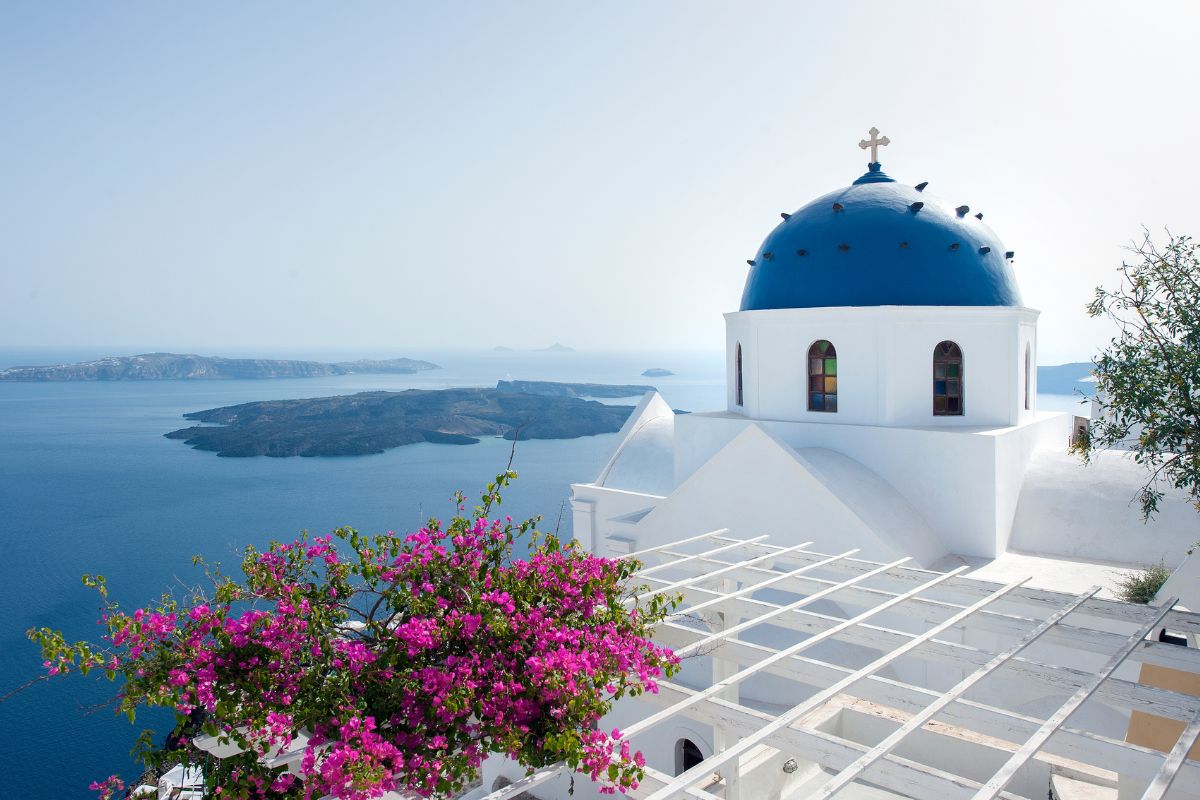 A classic Santorini scene featuring a whitewashed building with a blue-domed church, pink flowers in the foreground, and a stunning view of the Aegean Sea and nearby islands in the background.