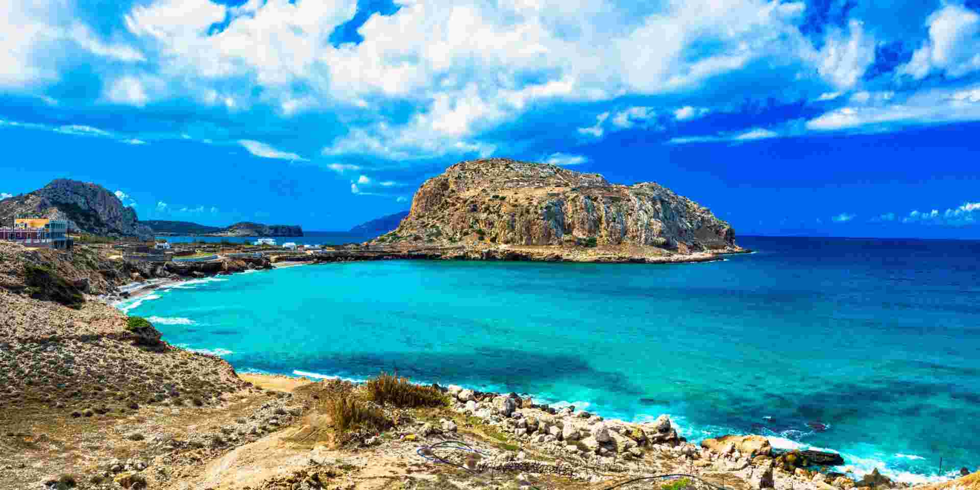 A scenic coastal view on Karpathos Island, showcasing turquoise waters, rocky terrain, and a prominent rocky hill under a partly cloudy sky. Karpathos Greece_Feature
