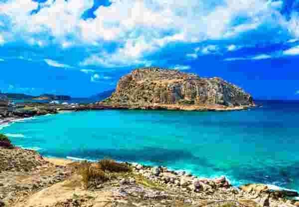 A scenic coastal view on Karpathos Island, showcasing turquoise waters, rocky terrain, and a prominent rocky hill under a partly cloudy sky. Karpathos Greece_Feature