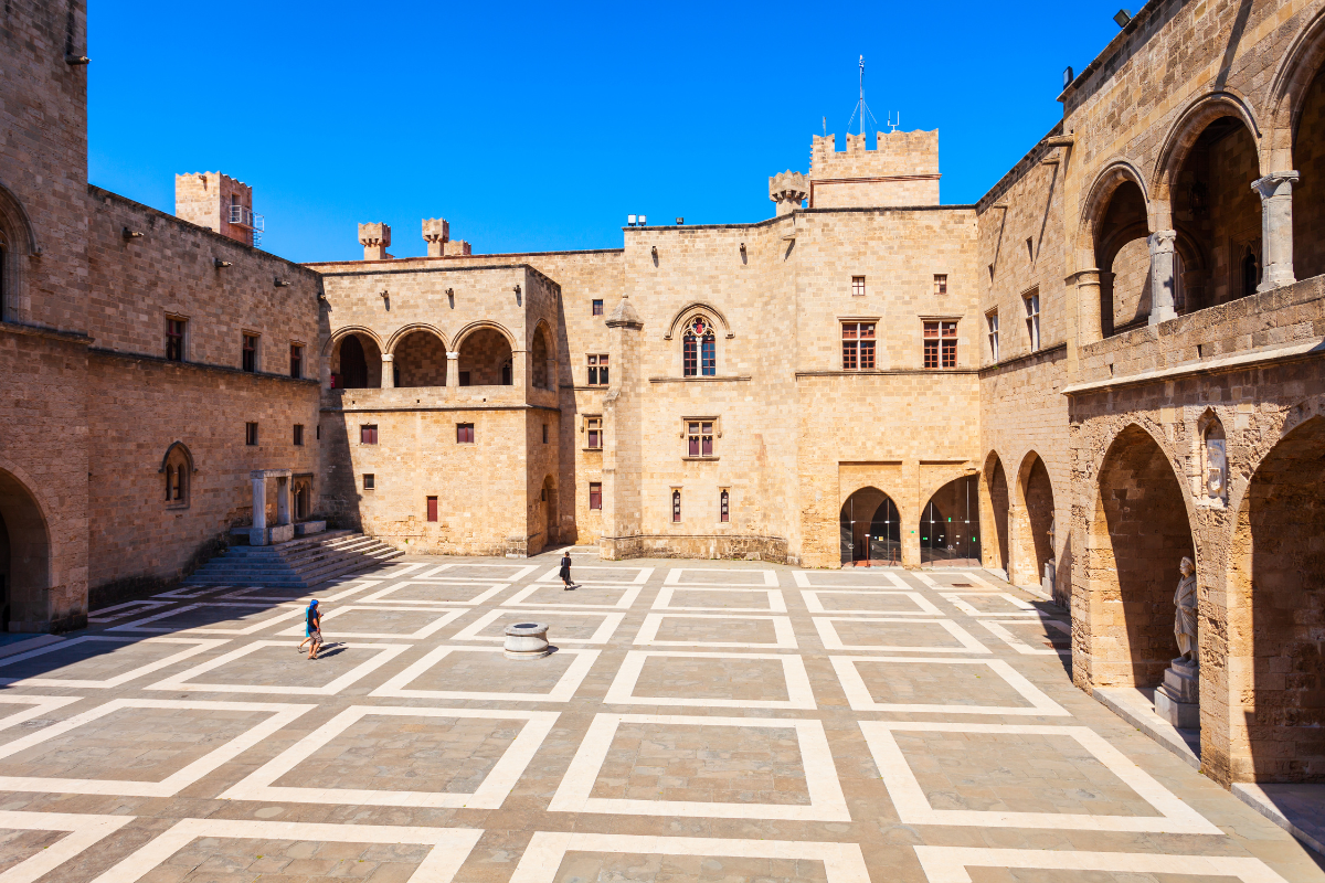 The image shows the courtyard of the Palace of the Grand Master in Rhodes, Greece, with its medieval stone architecture, large arches, and a patterned stone floor. A few people are walking around, highlighting the scale and grandeur of the historic building under a bright blue sky.