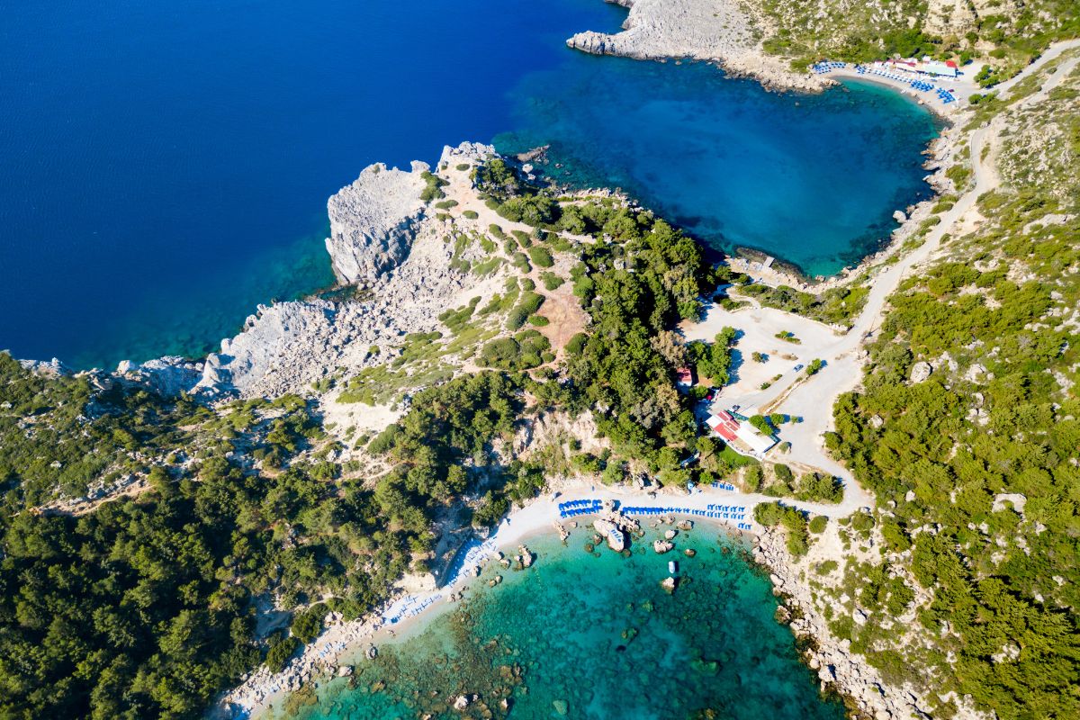 The image shows an aerial view of a beach in Rhodes, Greece, with clear turquoise waters, rocky coastlines, and lush greenery. A few beach umbrellas and sunbeds are visible along the shore, with small coves and inlets creating a scenic, secluded atmosphere.