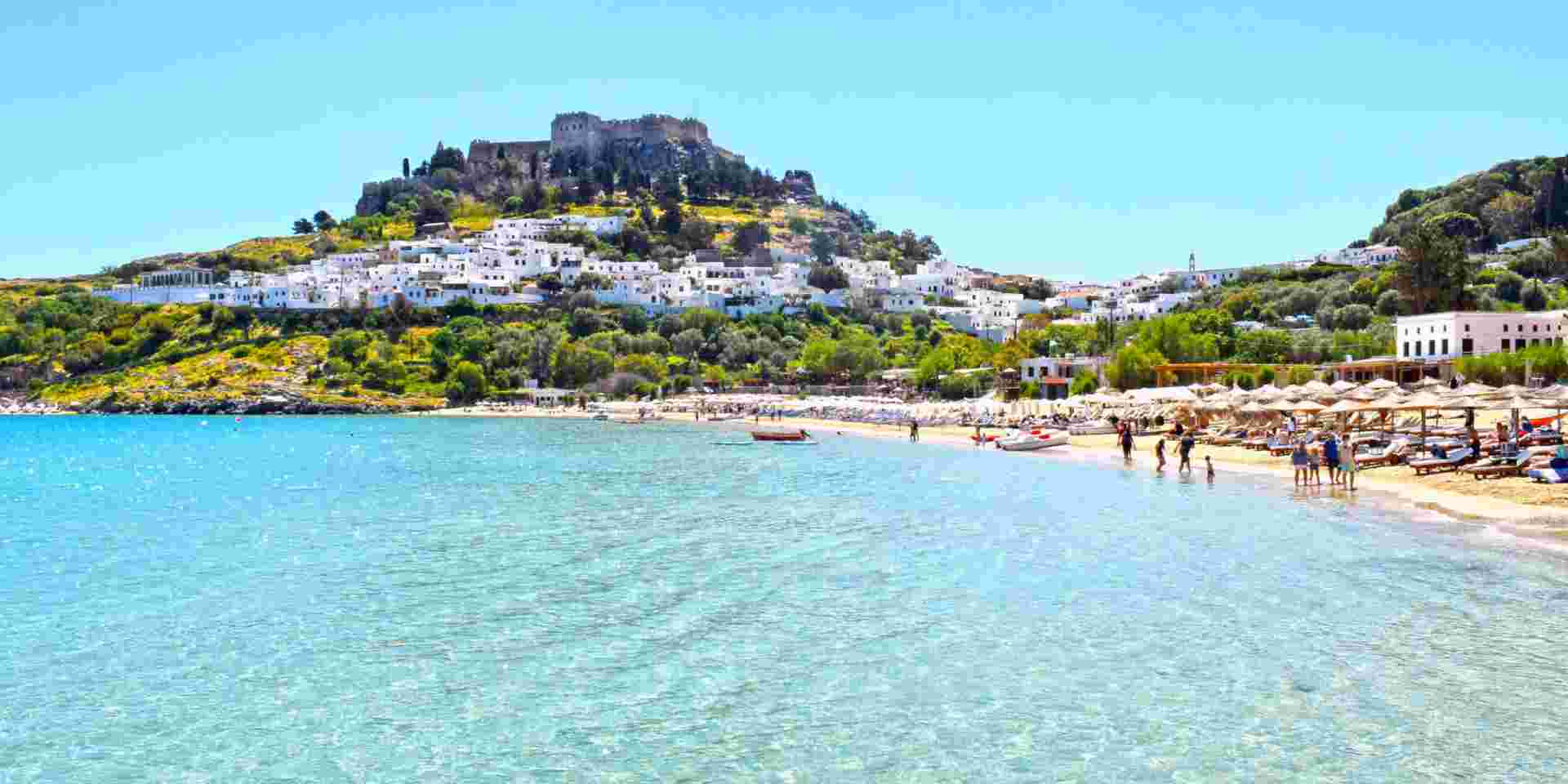 The image shows a beach in Rhodes, Greece, with clear blue waters and a sandy shoreline. In the background, there is a hilltop fortress surrounded by whitewashed buildings, with a few beachgoers enjoying the water and sun. rhodes greece_feature