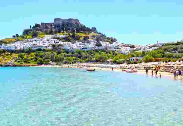 The image shows a beach in Rhodes, Greece, with clear blue waters and a sandy shoreline. In the background, there is a hilltop fortress surrounded by whitewashed buildings, with a few beachgoers enjoying the water and sun. rhodes greece_feature