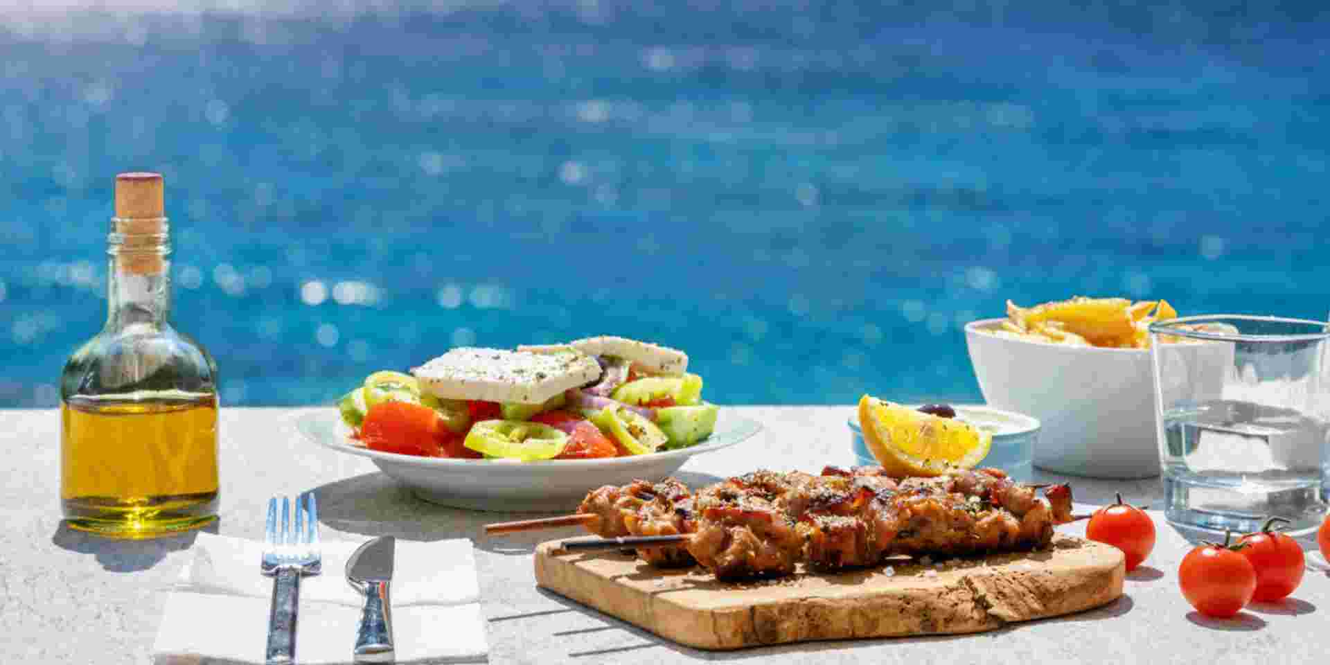 Greek food concept with farmers salad and souvlaki skewers in front of the sparkling, blue Aegean sea during summer time Greek food_feature