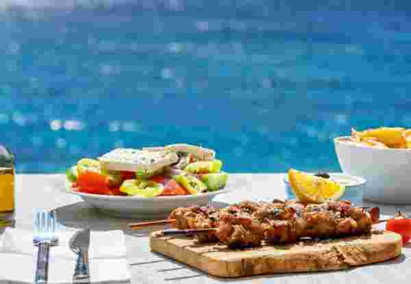 Greek food concept with farmers salad and souvlaki skewers in front of the sparkling, blue Aegean sea during summer time Greek food_feature