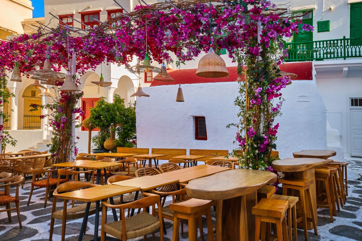 Cozy outdoor dining, cafe, restaurant, taverna in small Greek square, whitewashed chapel, vibrant pink bougainvillea in full blossom. Wooden tables, chairs, romantic dining, paradise destination, dream vacation.