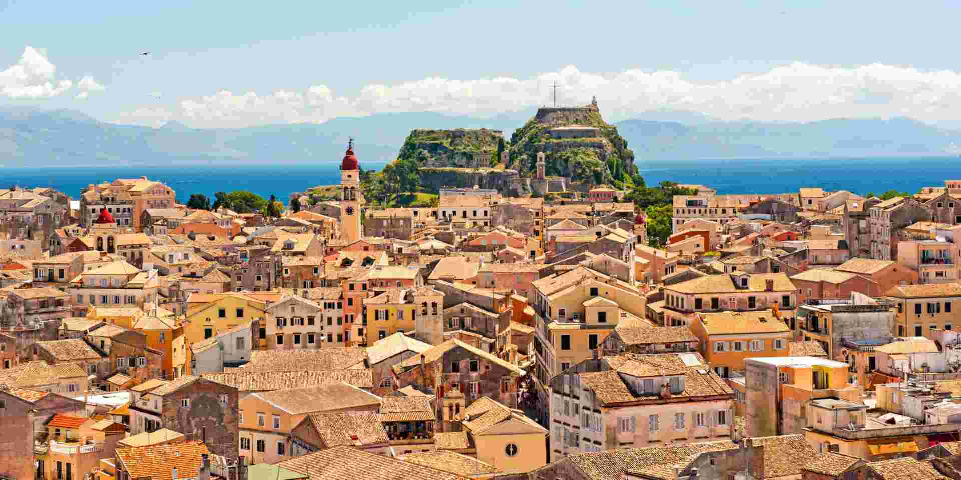A view of Old Town Corfu with its terracotta-roofed buildings, a prominent red-domed bell tower, and the Old Fortress in the background. The blue sea and distant mountains complete the scenic backdrop. corfu old town_monuments in greece_feature