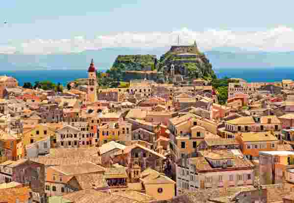 A view of Old Town Corfu with its terracotta-roofed buildings, a prominent red-domed bell tower, and the Old Fortress in the background. The blue sea and distant mountains complete the scenic backdrop. corfu old town_monuments in greece_feature