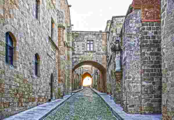 A cobblestone street in the medieval city of Rhodes, lined with stone buildings featuring arched windows and historic architectural details. An arched passageway leads to a bright opening in the distance. rhodes_monuments in greece