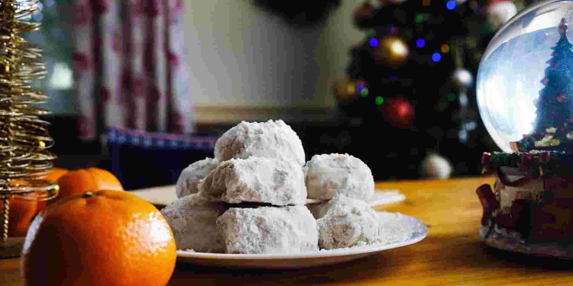 A plate of powdered sugar-coated kourabiedes, traditional Greek Christmas cookies, placed on a festive table with oranges and holiday decorations in the background. Greek Christmas Food_Feature