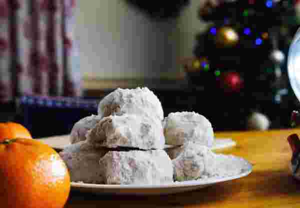 A plate of powdered sugar-coated kourabiedes, traditional Greek Christmas cookies, placed on a festive table with oranges and holiday decorations in the background. Greek Christmas Food_Feature