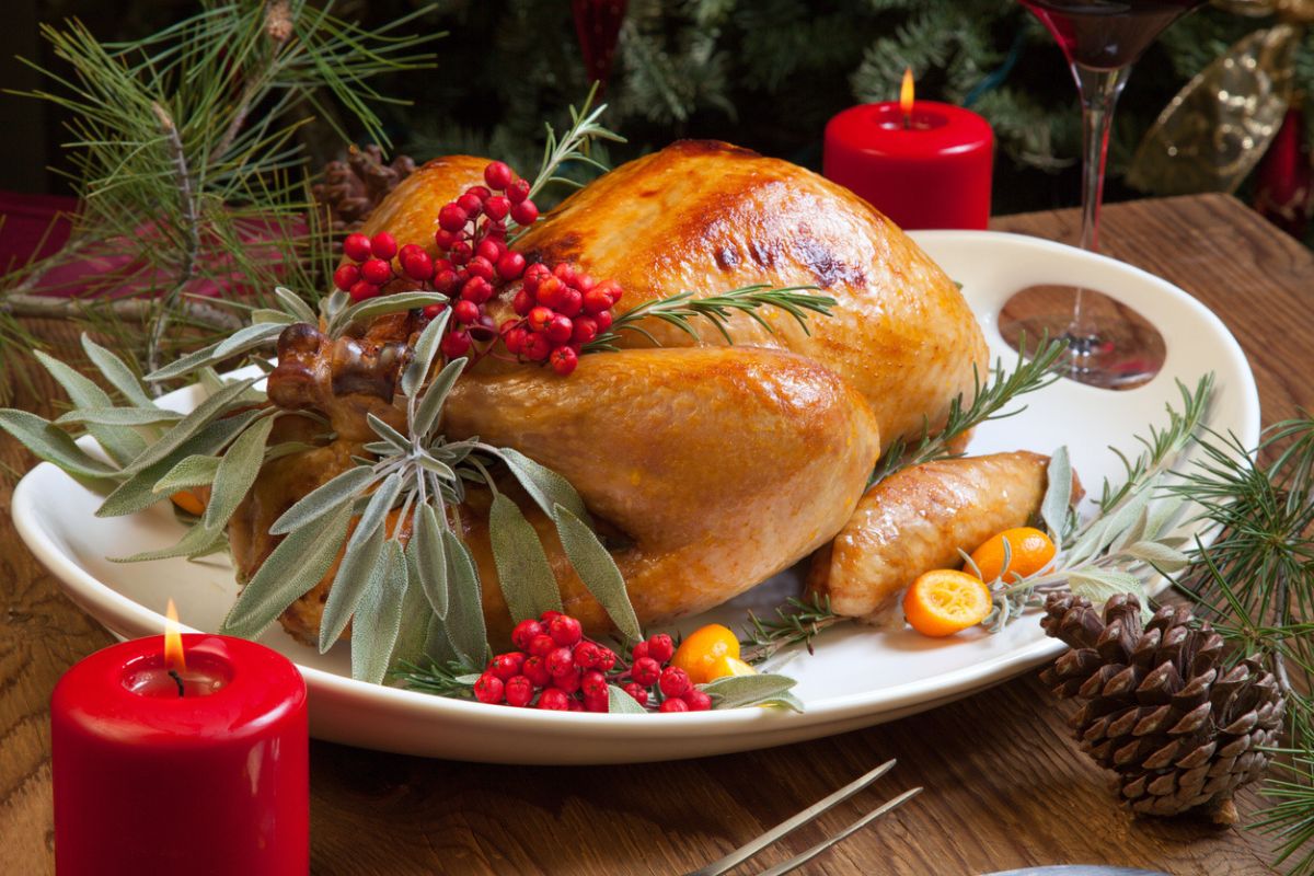 Roasted turkey garnished with sage, rosemary, and red berries in a tray prepared for Christmas dinner. Holiday table, candles and Christmas tree with ornaments.