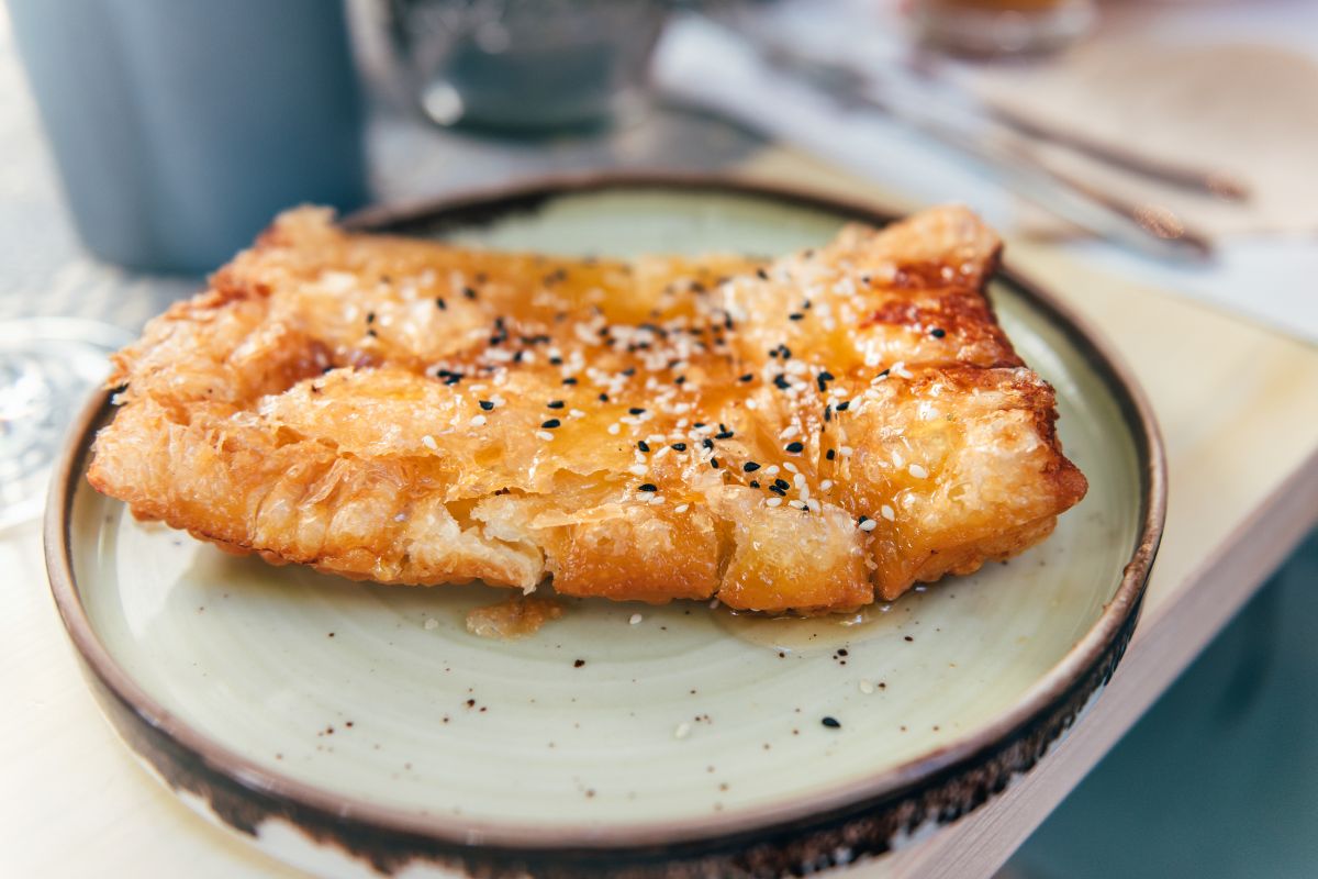 A close-up of a golden, flaky pastry topped with sesame seeds and drizzled with honey, with a savory feta cheese filling.