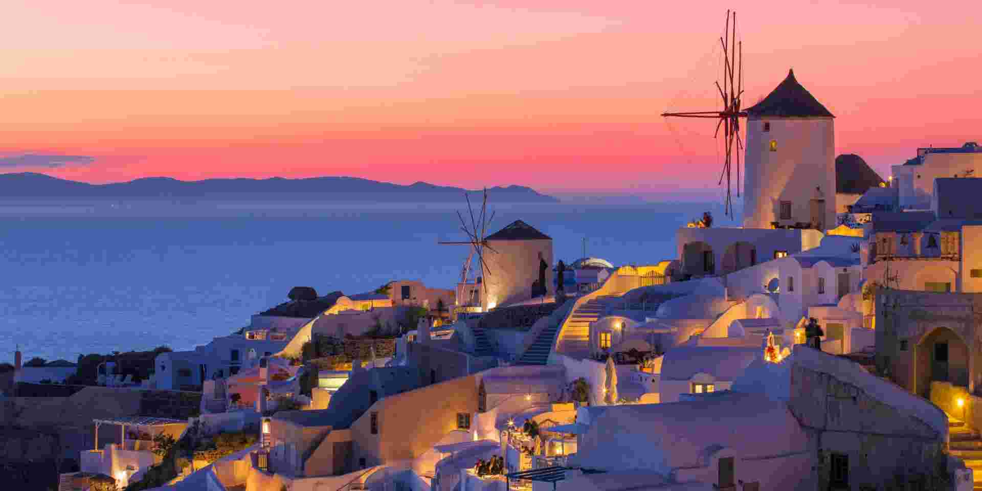 A scenic view of a Greek island at sunset, featuring traditional whitewashed buildings, windmills, and the sea blending into a vibrant, colorful sky. Santorini sunset_greece sunsets_feature