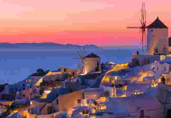 A scenic view of a Greek island at sunset, featuring traditional whitewashed buildings, windmills, and the sea blending into a vibrant, colorful sky. Santorini sunset_greece sunsets_feature