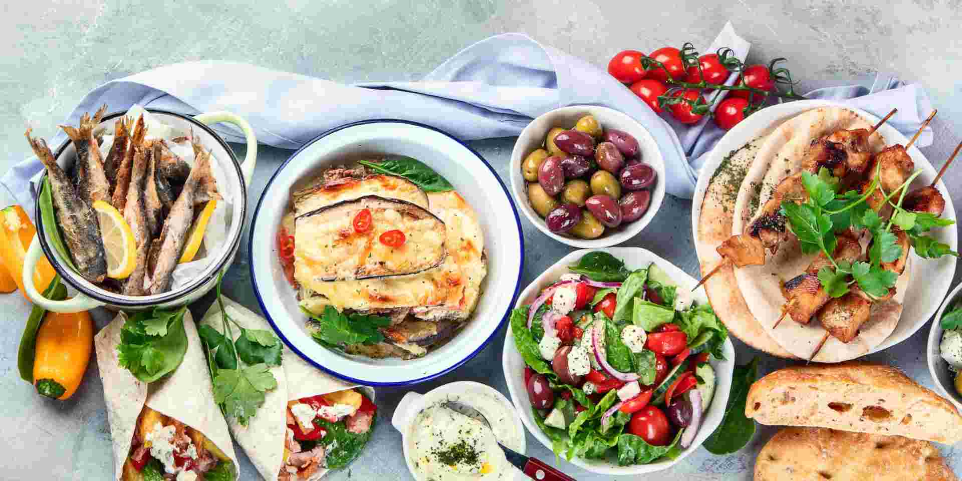 A spread of Greek foods, including grilled meats, fresh salad, olives, pita bread, fried fish, and baked moussaka, arranged on a table with colorful garnishes. greek foods_feature