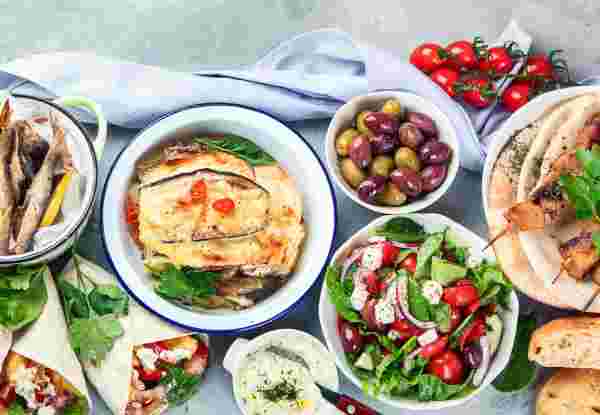 A spread of Greek foods, including grilled meats, fresh salad, olives, pita bread, fried fish, and baked moussaka, arranged on a table with colorful garnishes. greek foods_feature
