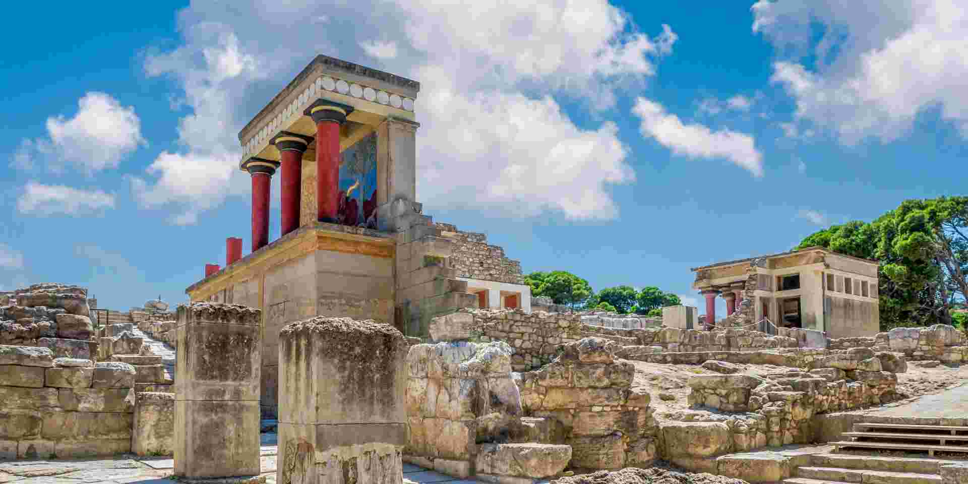 A partially restored section of the Knossos Palace complex, featuring red columns and stone ruins under a bright sky. knossos palace_historical sites in Greece_feature