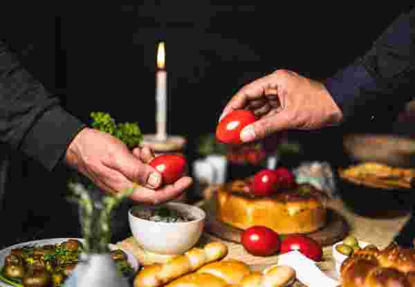Two hands holding red Easter eggs during a traditional Greek Easter celebration, with a table set with festive food, bread, and a lit candle in the background. Greek Easter Traditionsl