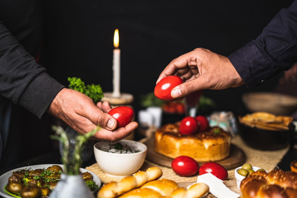 Two hands holding red Easter eggs during a traditional Greek Easter celebration, with a table set with festive food, bread, and a lit candle in the background. Greek Easter Traditionsl