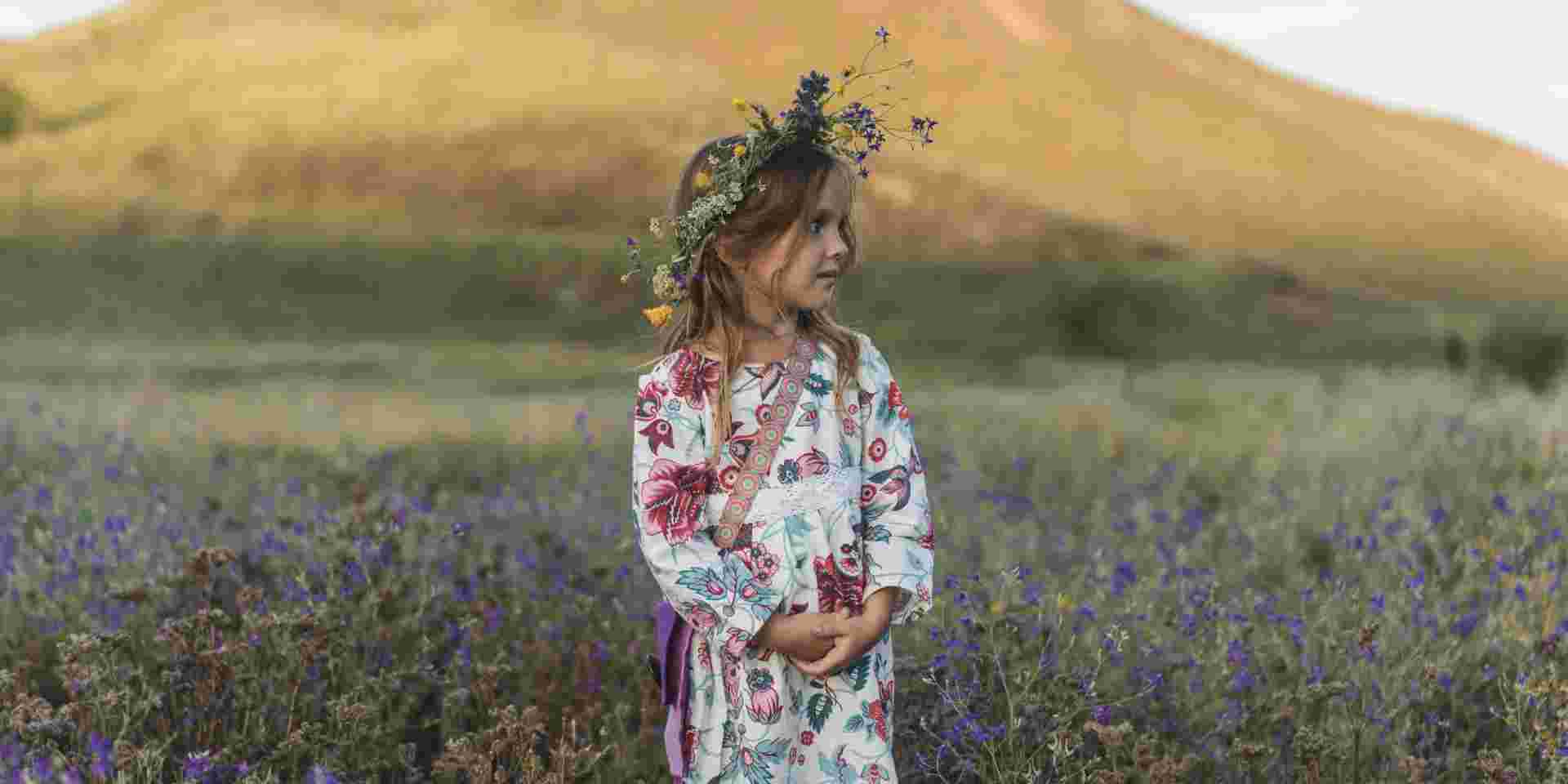 Young girl wearing a floral dress and flower crown standing in a spring field, representing May Day customs in Greece. may day customs in greece_feature