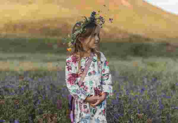 Young girl wearing a floral dress and flower crown standing in a spring field, representing May Day customs in Greece. may day customs in greece_feature