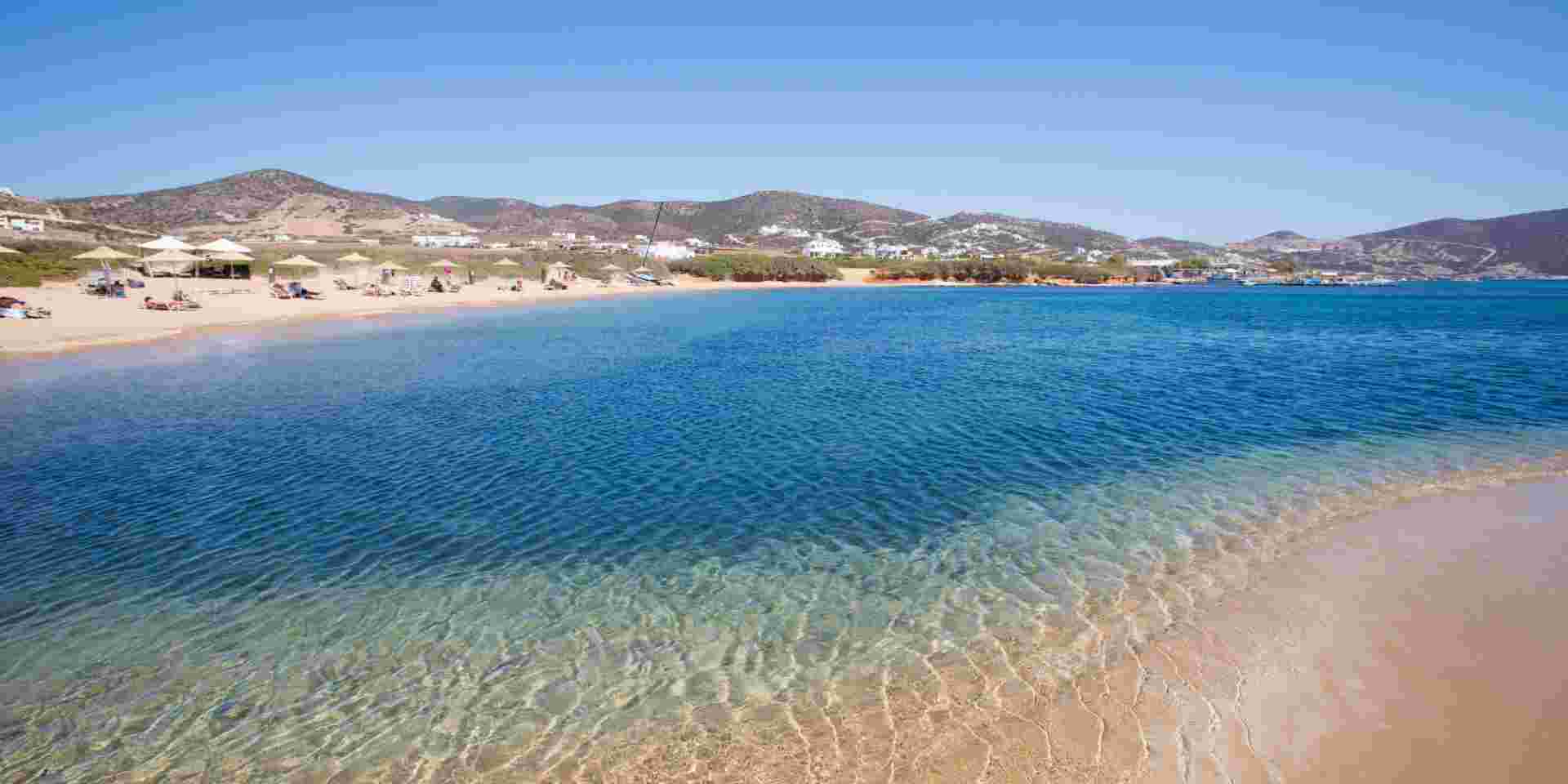 A tranquil beach in Antiparos with crystal-clear waters gently meeting the sandy shore, dotted with umbrellas and sun loungers, and hills in the background under a bright blue sky. antiparos greece_feature