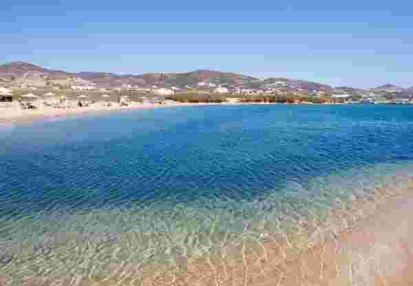 A tranquil beach in Antiparos with crystal-clear waters gently meeting the sandy shore, dotted with umbrellas and sun loungers, and hills in the background under a bright blue sky. antiparos greece_feature