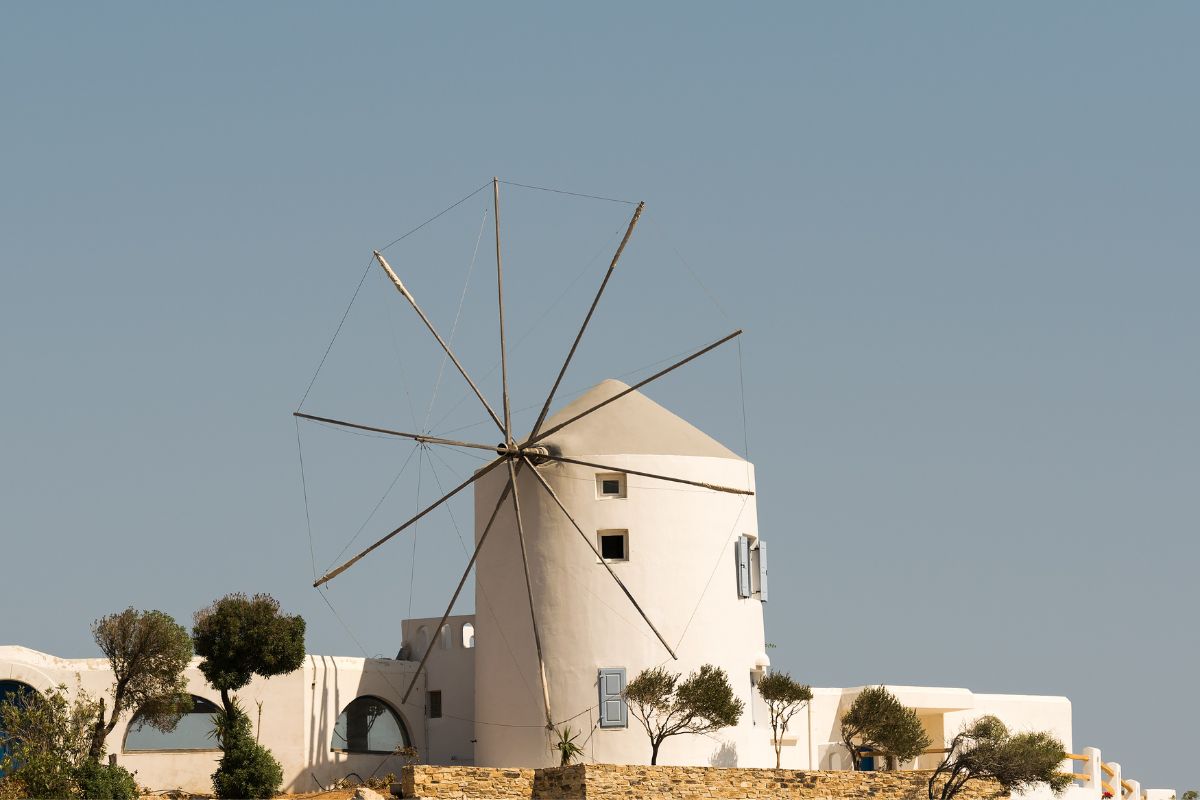 A traditional white windmill in Antiparos, standing against a clear blue sky, surrounded by small trees and minimal architecture, evoking a peaceful, timeless feel.