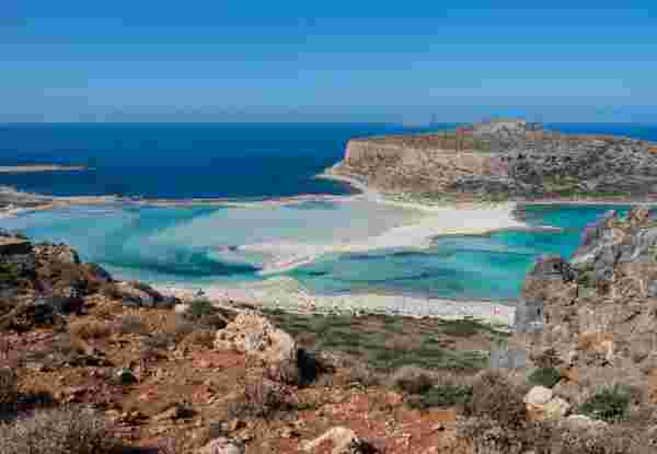 A scenic view of Balos Beach in Chania, featuring clear turquoise waters, white sandy shores, and a rugged hill in the background. A few boats are anchored near the shoreline, and people are scattered across the beach enjoying the sunny day. Balos beach_chania