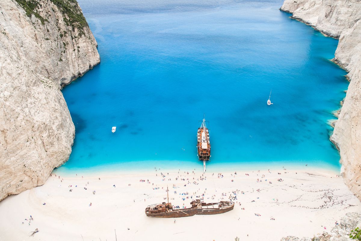 An aerial view of Navagio Beach in Zakynthos, featuring clear blue waters, white sandy shores, and a shipwreck on the beach surrounded by towering cliffs.