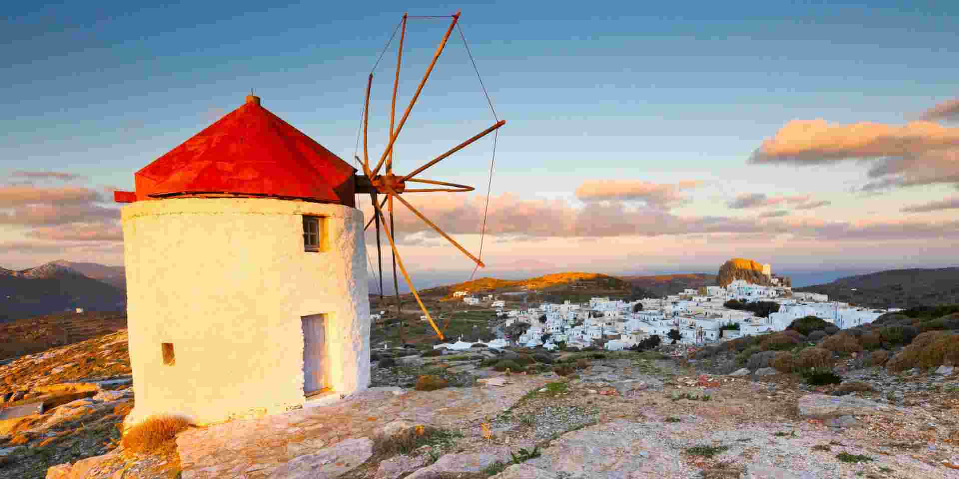 A picturesque view of Amorgos featuring a traditional white windmill with a red roof, overlooking a village of whitewashed houses under a soft, golden sunset. The rugged landscape stretches out in the background. Amorgos Greece_feature