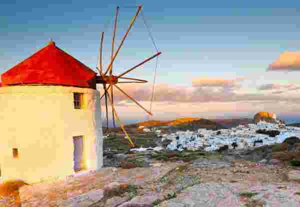 A picturesque view of Amorgos featuring a traditional white windmill with a red roof, overlooking a village of whitewashed houses under a soft, golden sunset. The rugged landscape stretches out in the background. Amorgos Greece_feature
