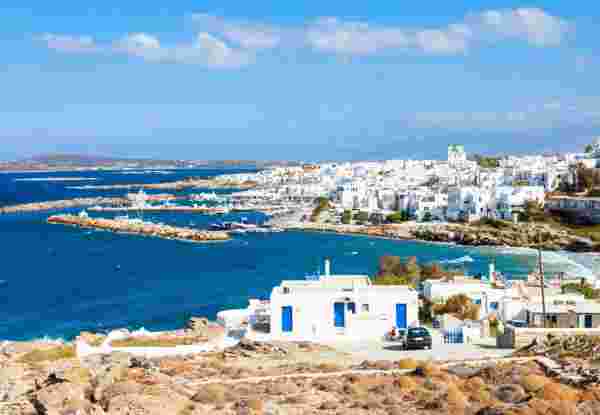 A view of Paros Island featuring whitewashed buildings along the coastline, with the deep blue Aegean Sea and clear skies in the background. Paros Island_greece in july
