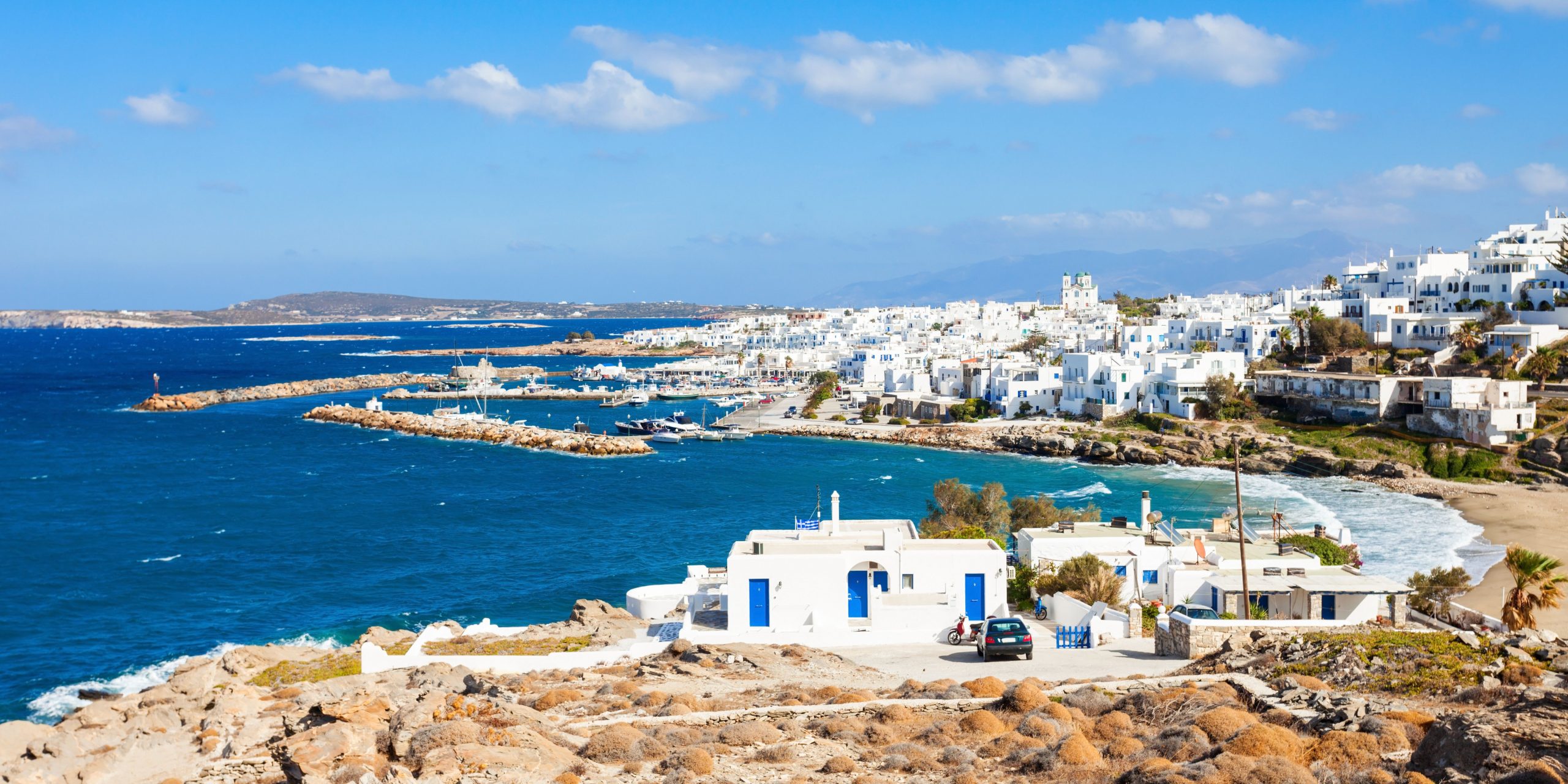 A view of Paros Island featuring whitewashed buildings along the coastline, with the deep blue Aegean Sea and clear skies in the background.