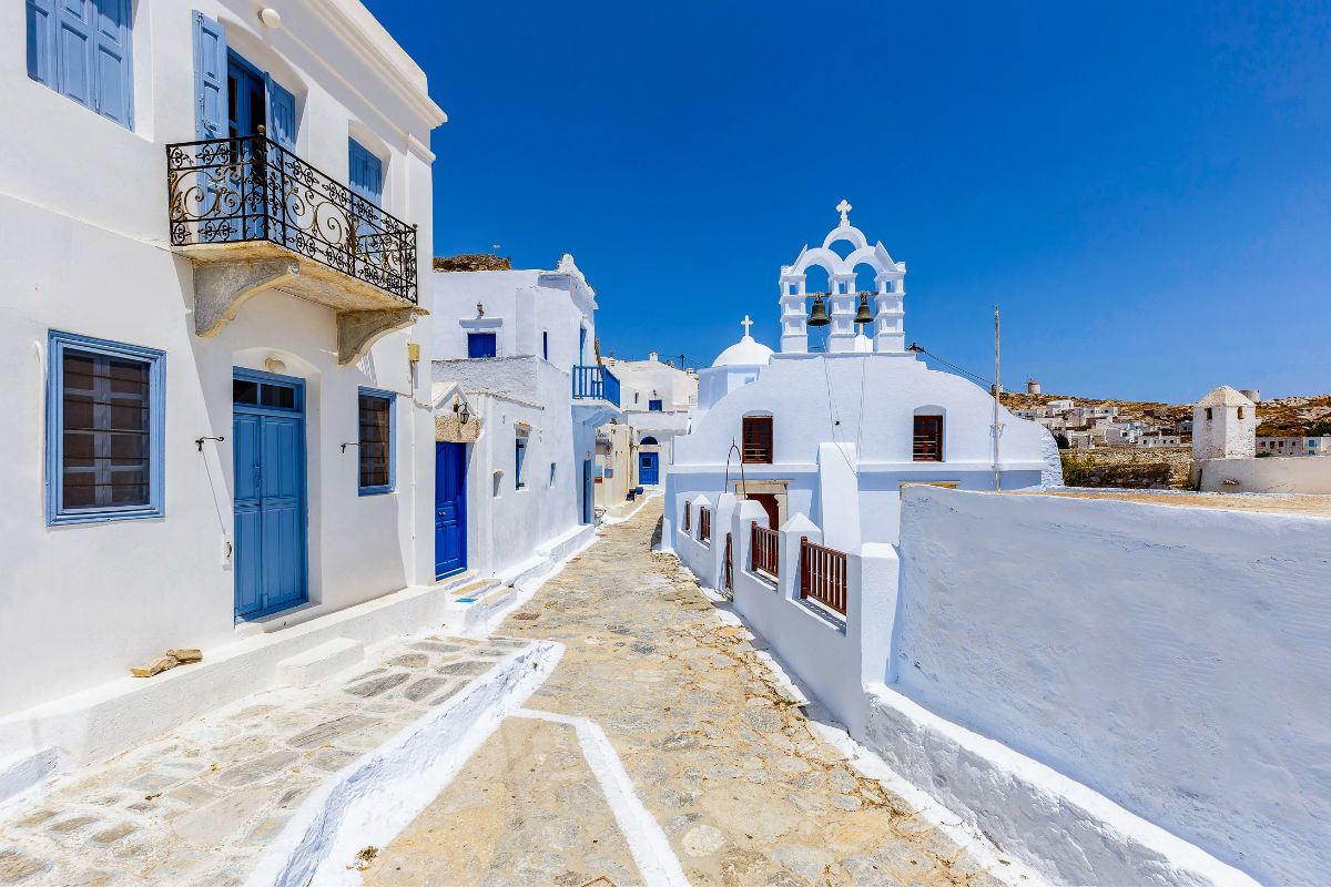 A narrow, sunlit street in Amorgos lined with traditional whitewashed buildings, blue doors, and windows. In the background, a white church with bell towers stands under a bright blue sky.