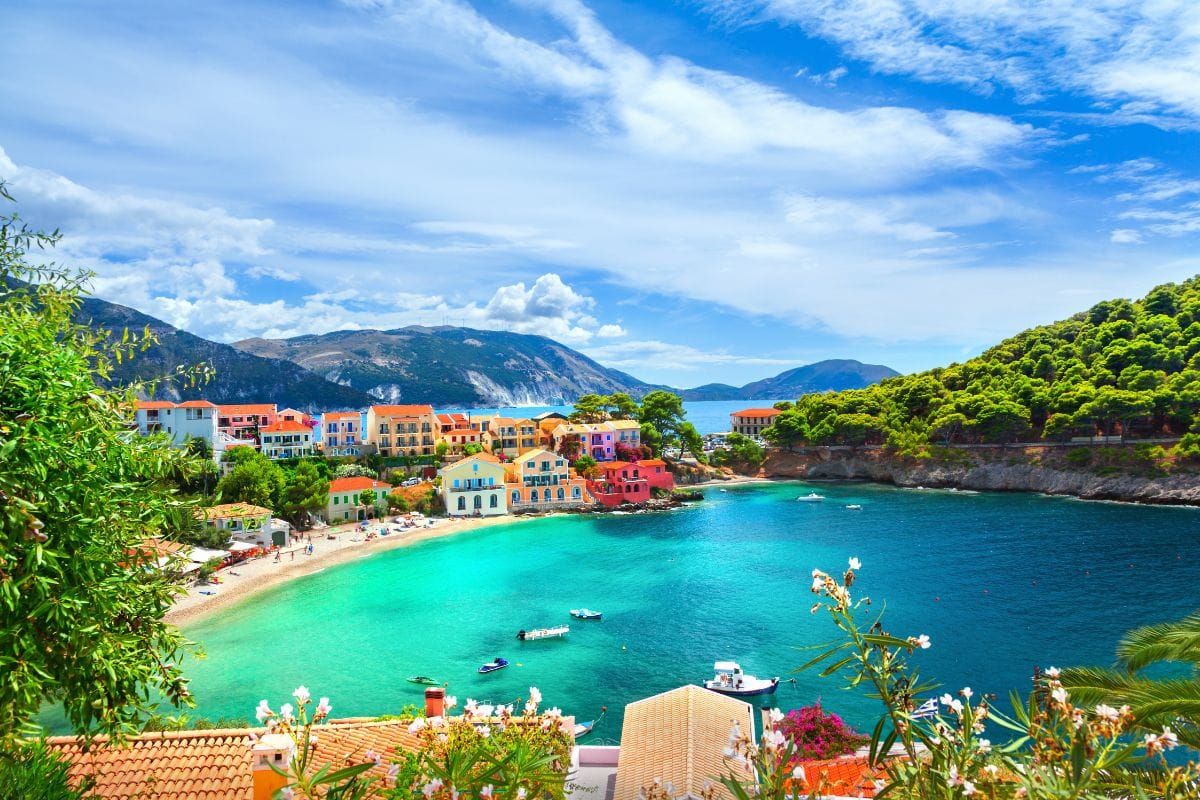 A picturesque view of Assos Village in Kefalonia, featuring colorful houses along a turquoise bay surrounded by lush greenery and mountains in the background.