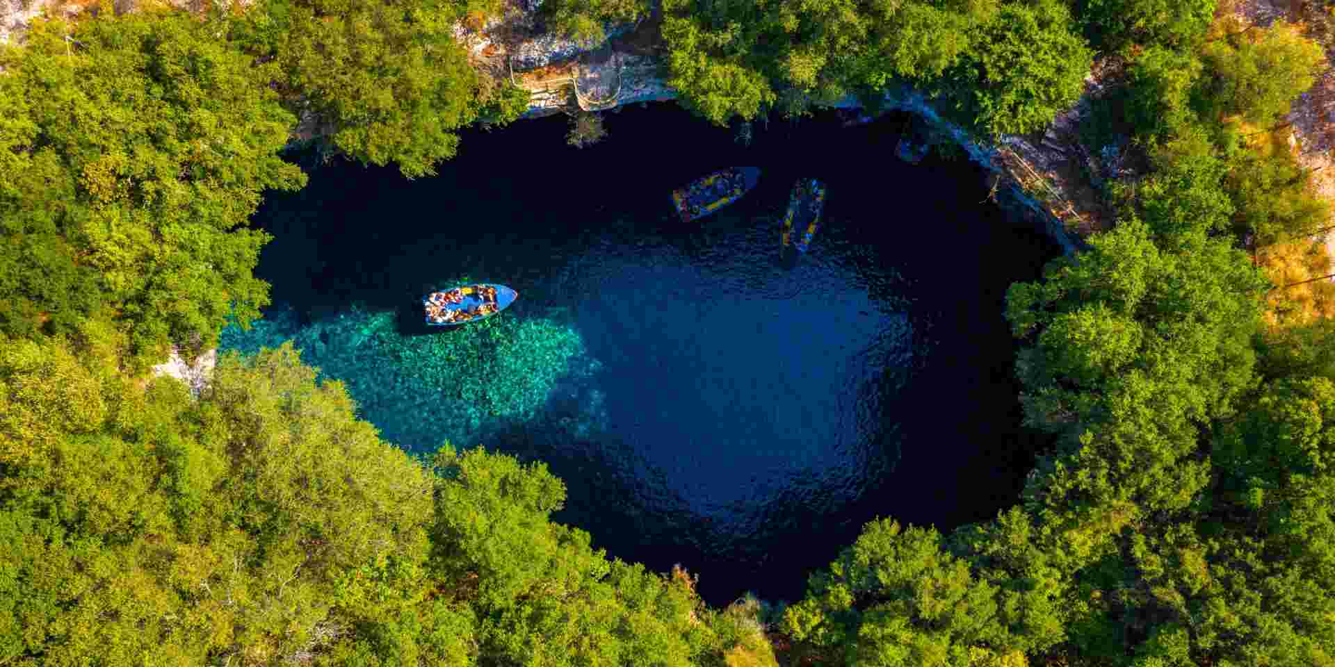 Aerial view of Melissani Lake in Kefalonia, showing boats with tourists on the clear blue water surrounded by lush green trees. melissani cave_things to do in kefalonia_feature