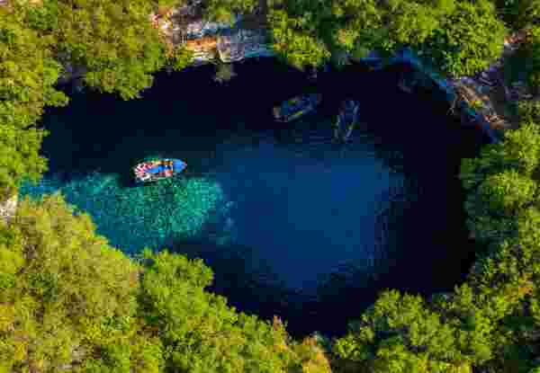 Aerial view of Melissani Lake in Kefalonia, showing boats with tourists on the clear blue water surrounded by lush green trees. melissani cave_things to do in kefalonia_feature