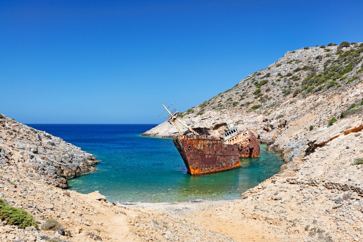 A view of a rusted shipwreck resting in a small cove with turquoise waters, surrounded by rocky hills, under a bright blue sky on Amorgos Island.