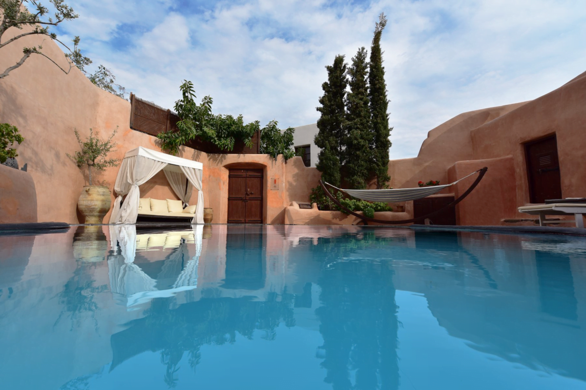 A serene pool area with a canopied daybed and hammock, surrounded by terracotta walls and greenery under a clear sky.