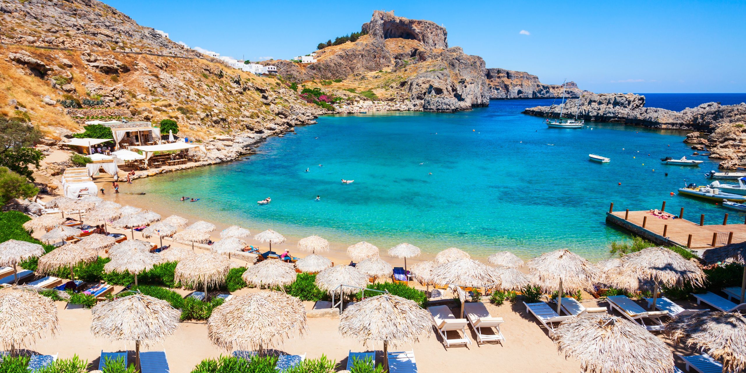 A picturesque beach cove in Rhodes with turquoise waters, thatched umbrellas, sunbeds, and a few boats docked nearby.