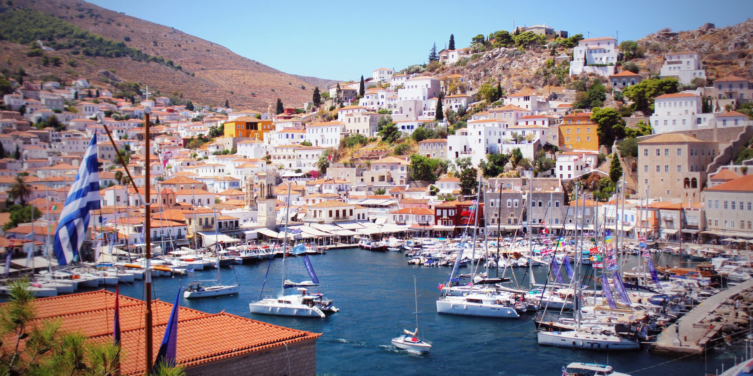 A vibrant harbor in Hydra with white-washed buildings, boats docked in the marina, and a hillside in the background._Best greek islands to visit in september
