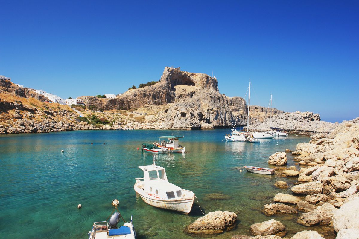 A cove with boats and clear blue water, overlooked by a rocky outcrop and buildings.