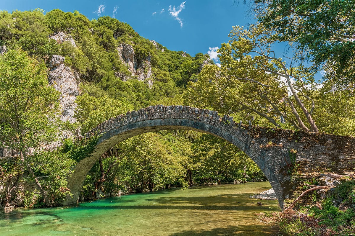 A stone arch bridge over a clear, green river surrounded by lush trees and rocky hills in Zagori._Explore Greece in October 2024: Your Ultimate Guide