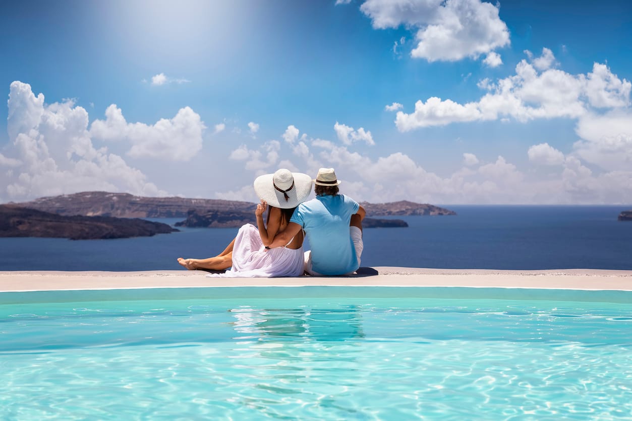 A elegant couple in summer clothes sits by the pool and enjoys the view to the mediterranean sea in Greece during their summer holidays