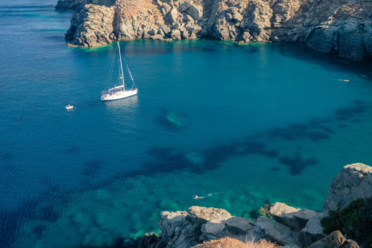 A sailboat anchored in a clear, blue bay surrounded by rocky cliffs, with a small dinghy and a swimmer nearby.